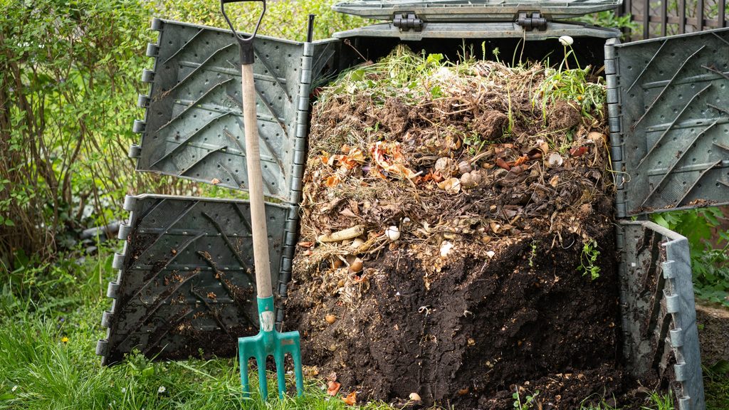 enclosed outdoor compost bin with pitchfork leaning against it in a backyard garden