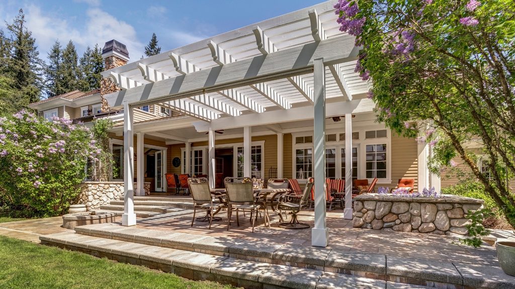 large white pergola attached to house over a stone patio with outdoor dining furniture