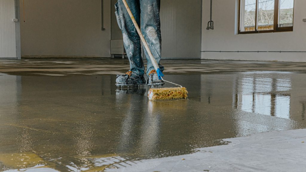 Person spreading epoxy coating across a concrete garage floor with a long-handled scrub brush