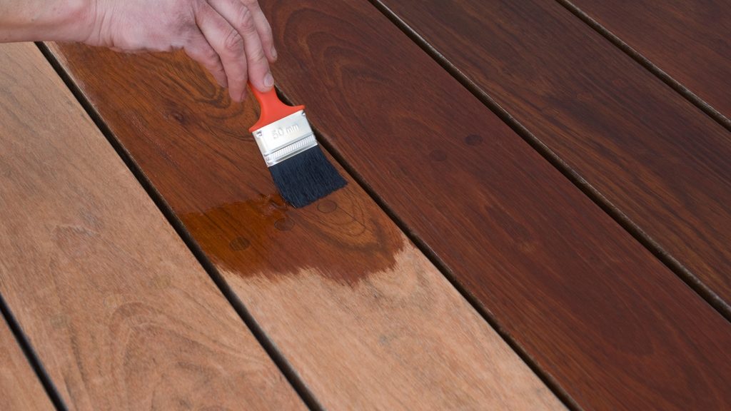 Hand using a brush to apply dark wood stain to deck boards along the grain