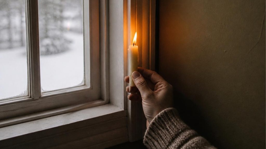Hand holding a lit candle near a window frame to detect a draft, with a snowy winter scene visible outside