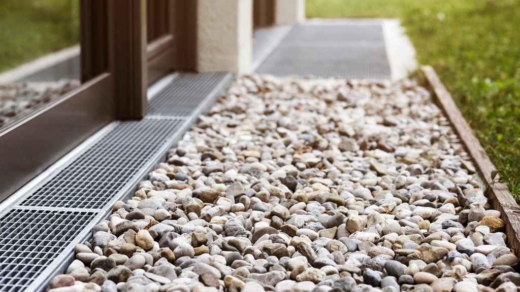 Channel drain with metal grate and river rock gravel along a home foundation