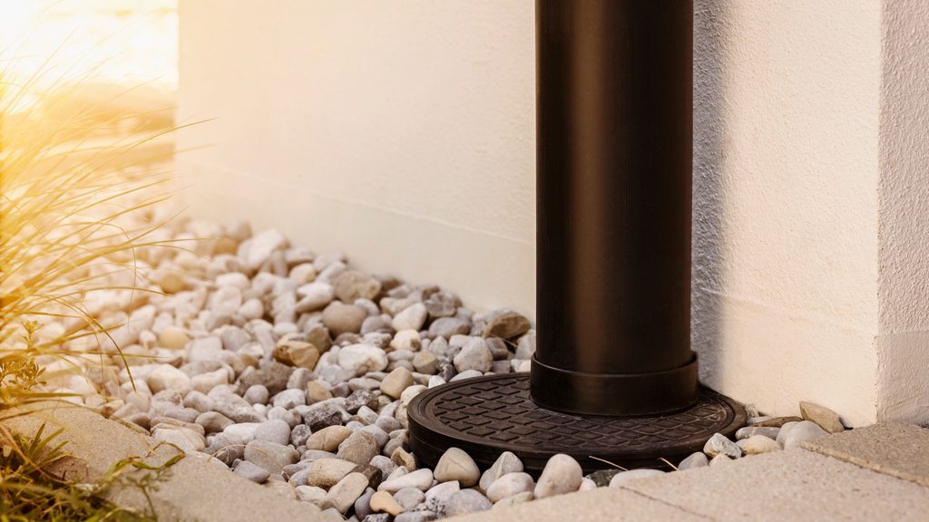 Downspout connected to an underground drain outlet surrounded by gravel and pavers near a home foundation