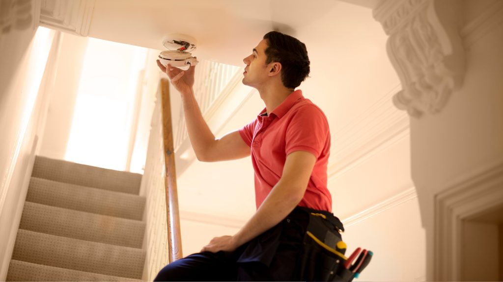 Man on a ladder installing a smoke detector on the ceiling above a staircase