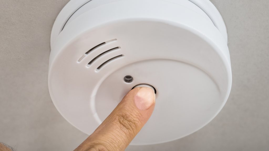 Finger pressing the test button on a white smoke detector mounted to a ceiling