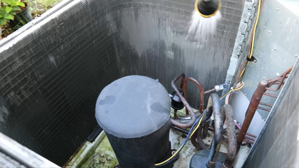 Garden hose rinsing the inside of an open AC condenser unit after coil cleaner application