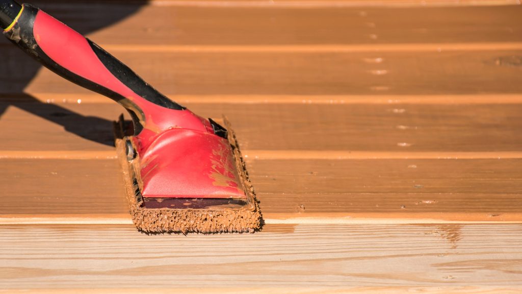 Red roller applicator spreading wood stain across deck boards