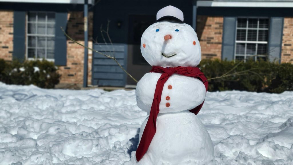 Snowman in a snowy front yard in front of a brick home — a reminder to winterize your sprinkler system before the first hard freeze