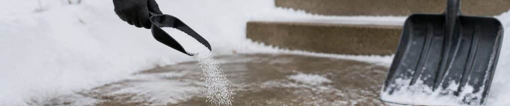 Gloved hand scattering ice melt from a scoop onto an icy concrete walkway, with a snow shovel nearby, choosing the best ice melt for concrete