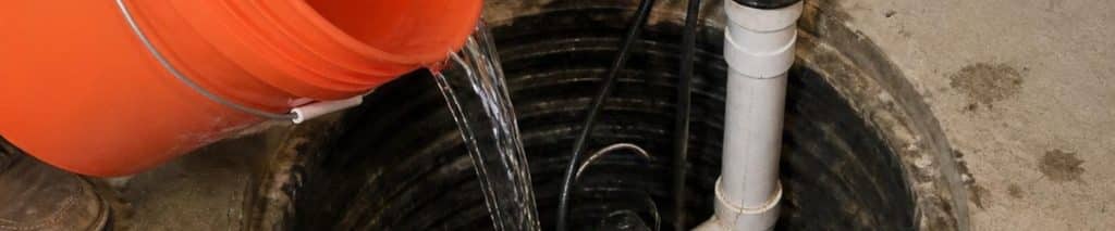 Pouring a bucket of water into a basement sump pit to test a sump pump, with the check valve on the discharge pipe and a flashlight nearby. How to test a sump pump