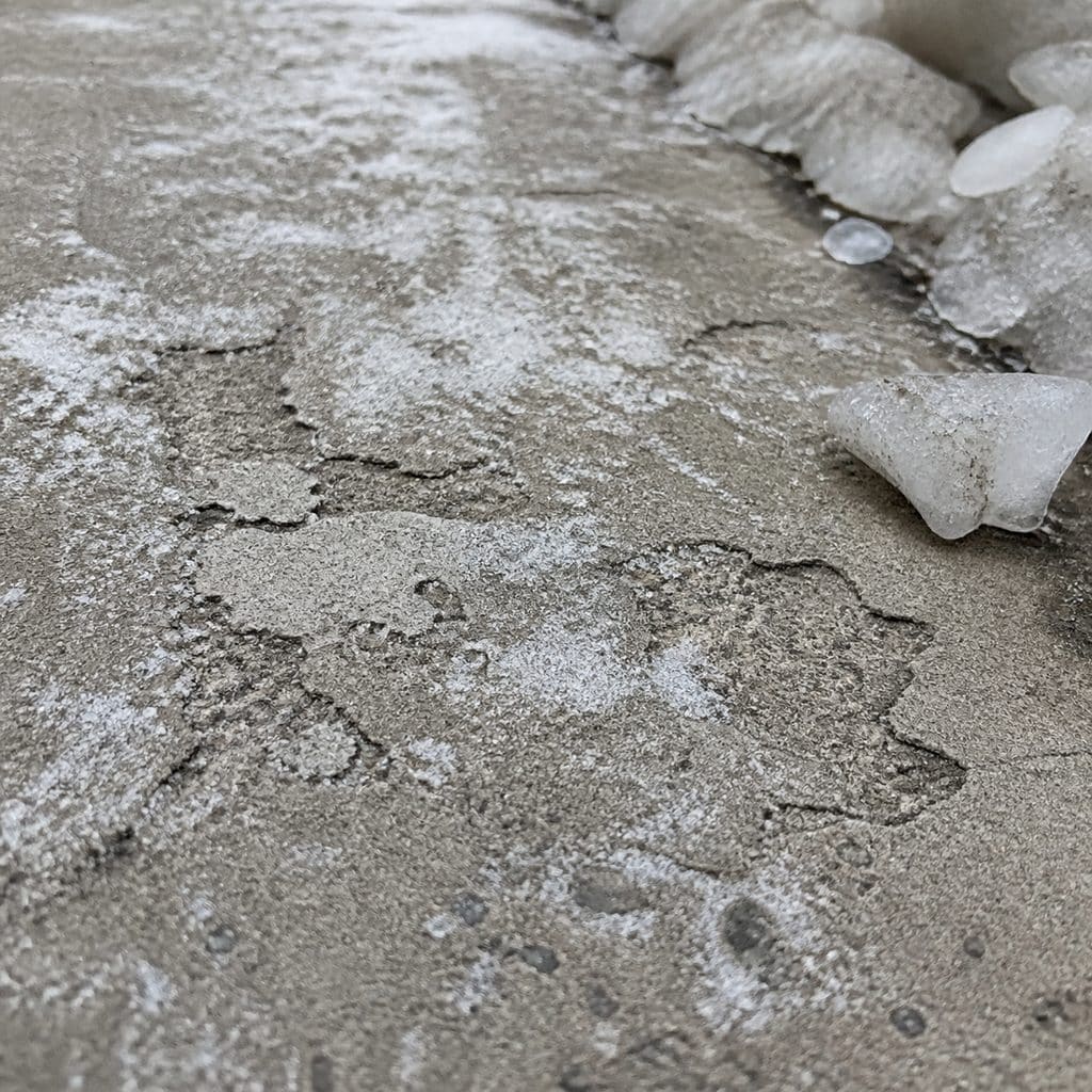 Concrete surface flaking and pitting from rock salt damage, with white salt residue and chunks of ice