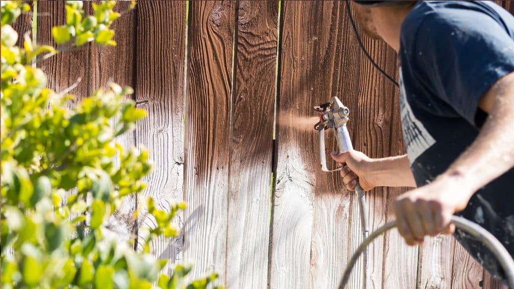 Spraying stain onto a weathered gray wood fence with an airless sprayer