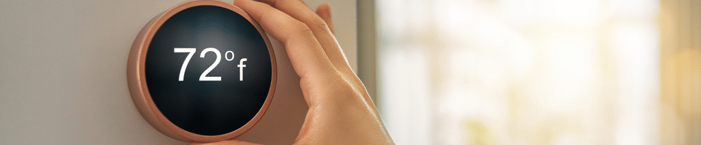Hand turning a round smart thermostat to adjust the temperature in a sunny living room. Thermostat savings calculator